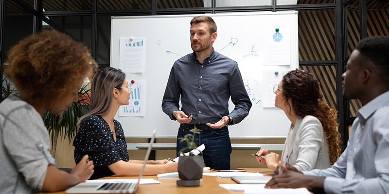 Confident businessman stand hold team business meeting in boardroom talk with multiethnic employees, Caucasian male presenter or coach speak make presentation brainstorm with businesspeople at meeting